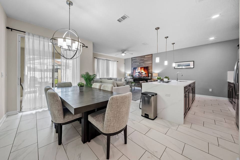 Our spacious dining area boasts a sleek wooden table with plush, light-colored chairs, ideal for gatherings. Large windows let in natural light, creating a warm, inviting atmosphere complemented by stylish pendant lighting.