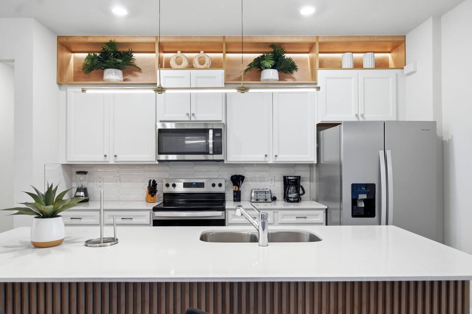 Welcome to this stylish kitchen with sleek white cabinetry and a spacious island, creating a bright, airy feel. Polished countertops, stainless steel appliances, and decorative shelves with greenery add to the inviting atmosphere.