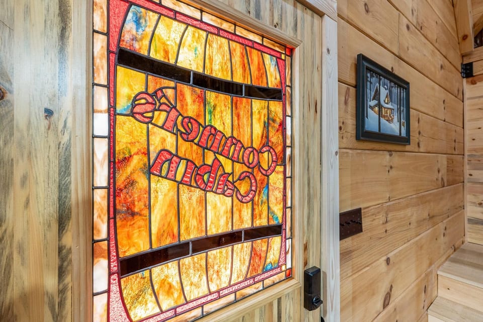 Entry door with amber-hued stained glass panel reading "Conner's Cabin" set in light pine walls with framed winter cabin art.