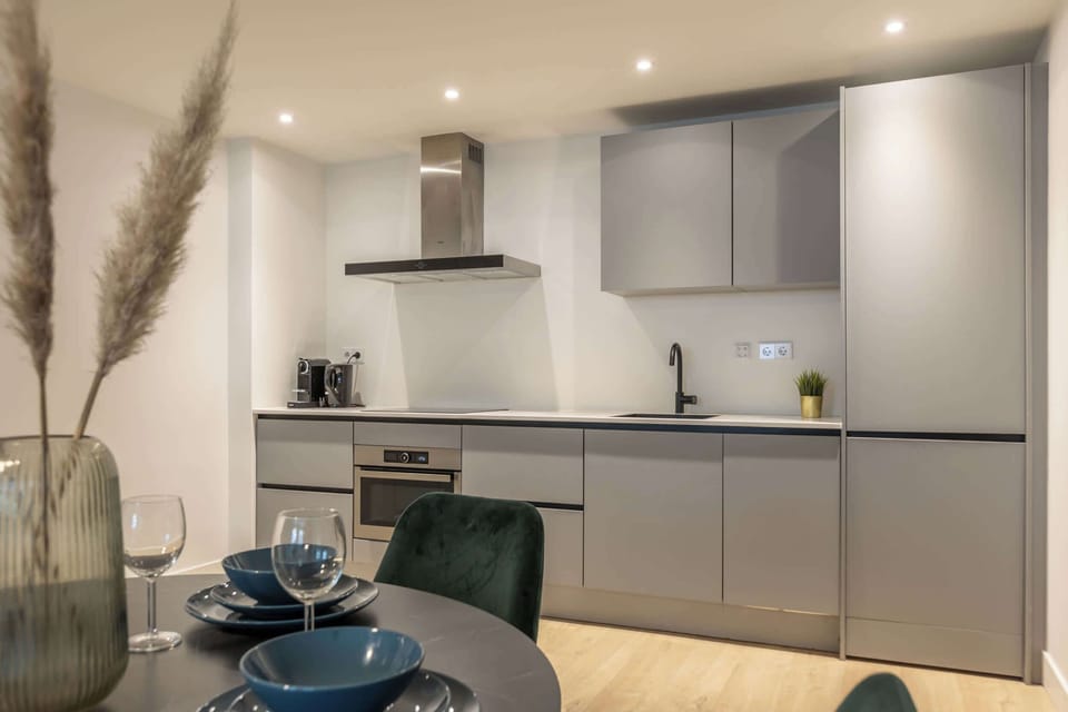 The fully equipped kitchen is integrated along the back wall and finished in matte grey cabinetry. It includes an oven, induction cooktop, extractor hood, fridge, and coffee machine.