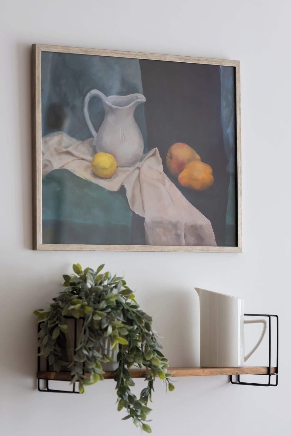 A decorative still-life painting hangs on the wall, depicting a pitcher and fruit in a classic style. Below it, a small wooden shelf holds a trailing green plant, adding a natural touch to the white wall.