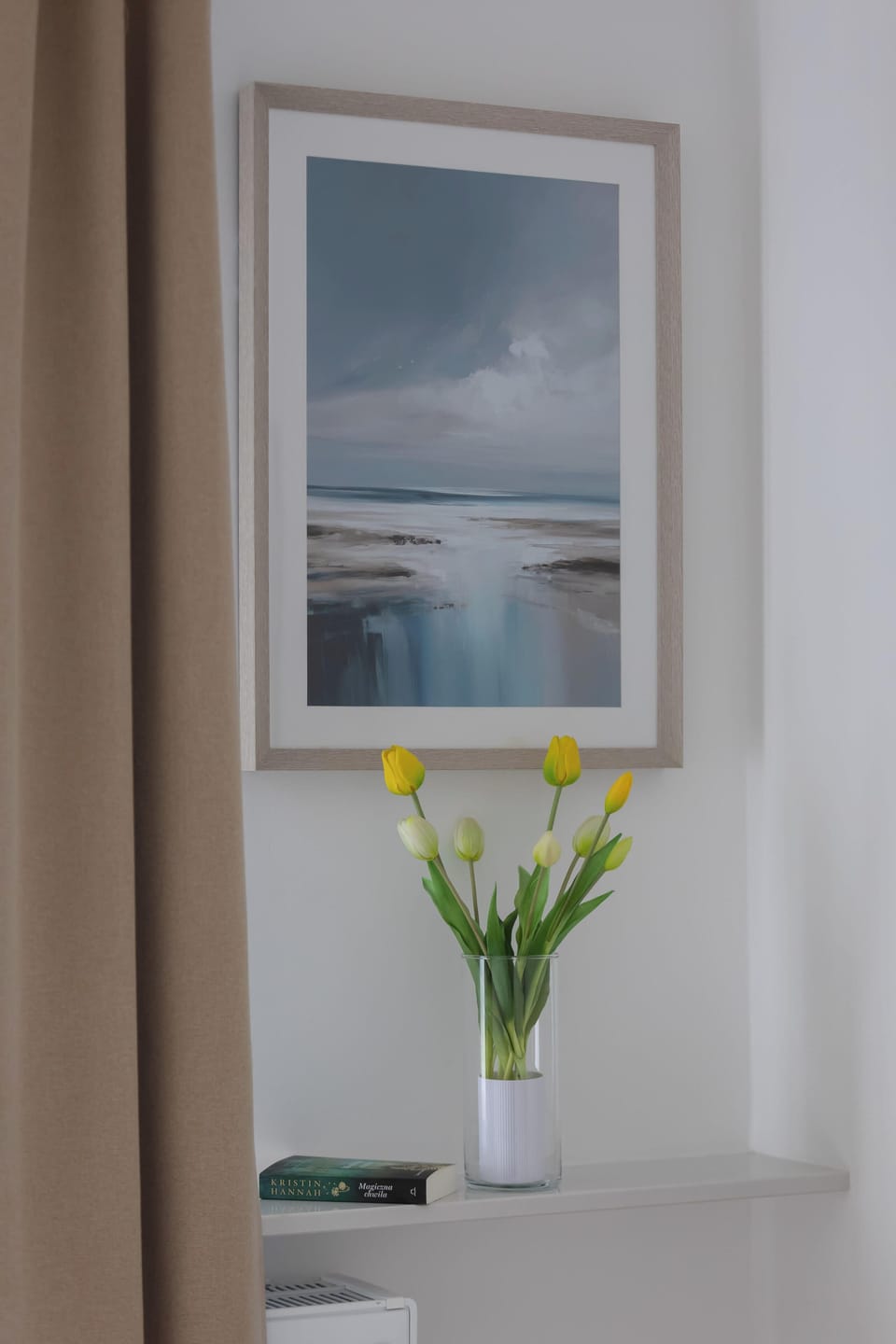 A vertical decorative shot shows a framed landscape painting hung above a small vase of yellow tulips. This arrangement sits on a narrow white shelf, adding a bright and fresh detail to the room.