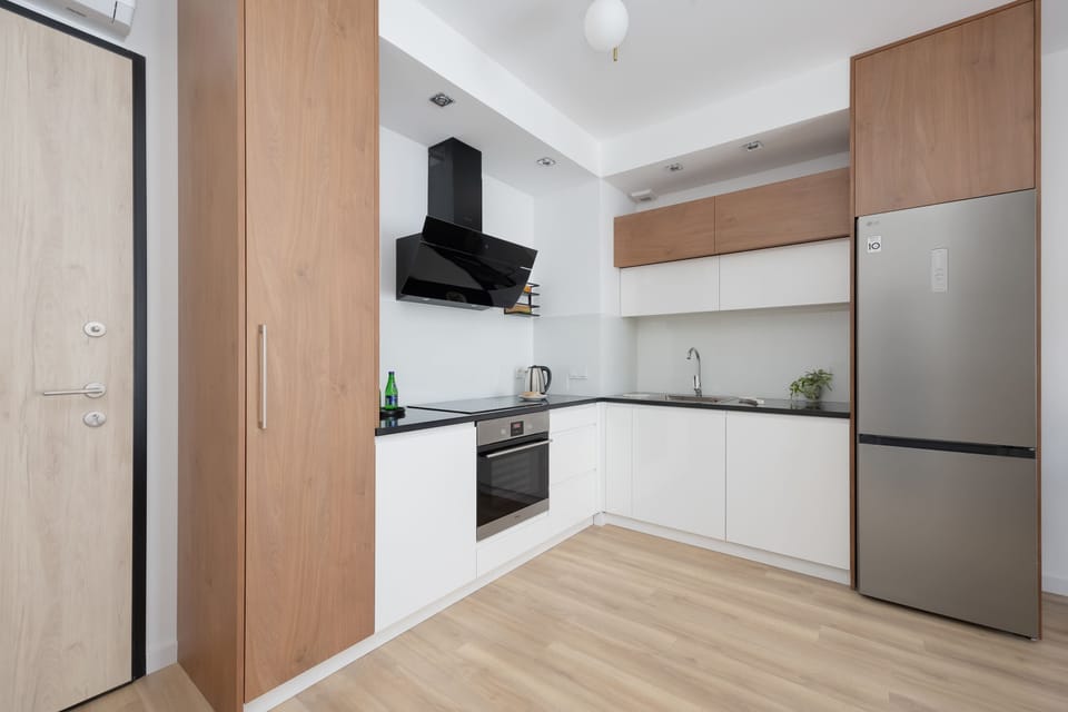 A modern kitchenette showcases clean white cabinetry paired with warm wood-toned accents. It is fully equipped with an oven, stovetop, and a large stainless steel refrigerator, all set on light-colored flooring.