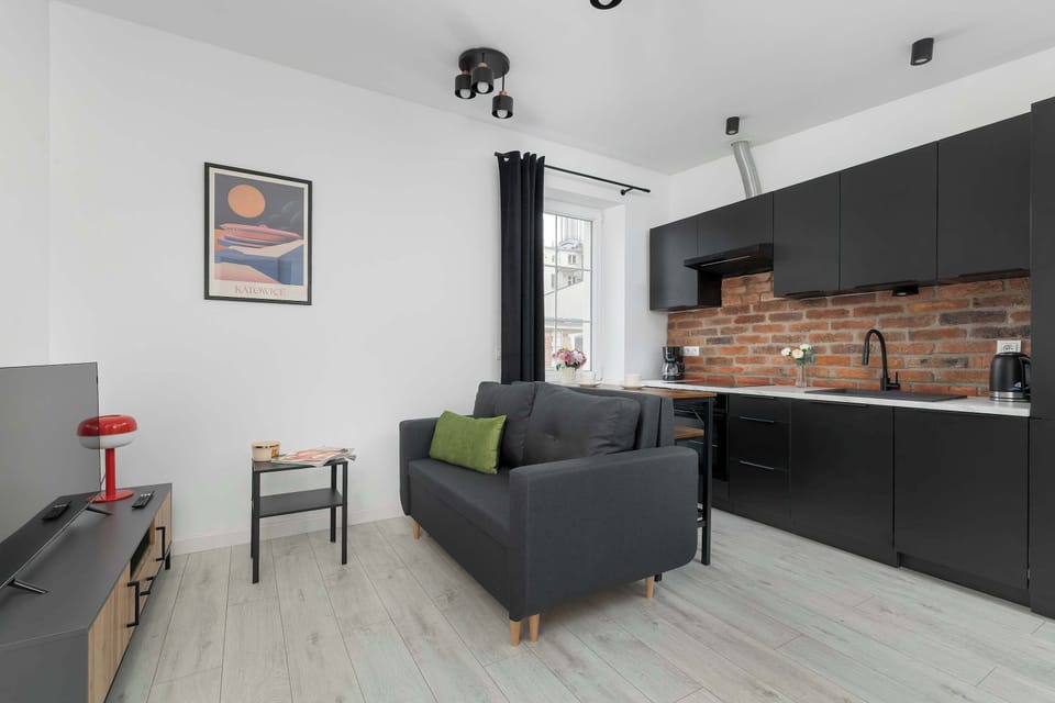 Bright modern living room with a gray sofa, white walls, and a stylish exposed brick kitchen in the background.