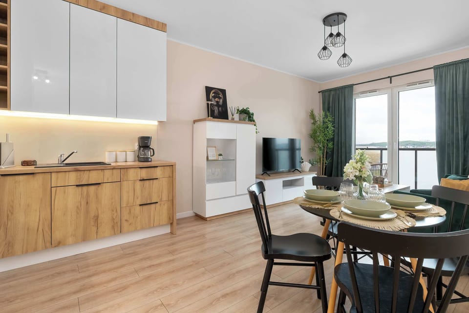 The open-plan layout connects the wooden-themed kitchen directly to the dining and living areas. Black chairs provide a sharp contrast to the light wood flooring and neutral wall colors.