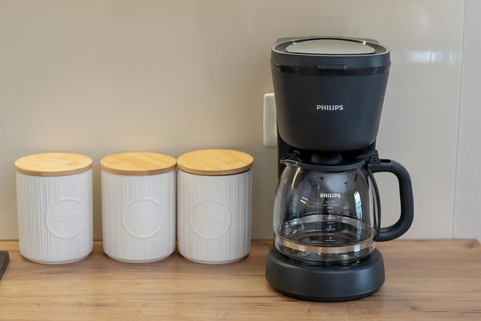 Three white ceramic canisters with wooden lids stand next to a black drip coffee machine on the kitchen counter. This organized station makes the kitchen feel practical and well-equipped.