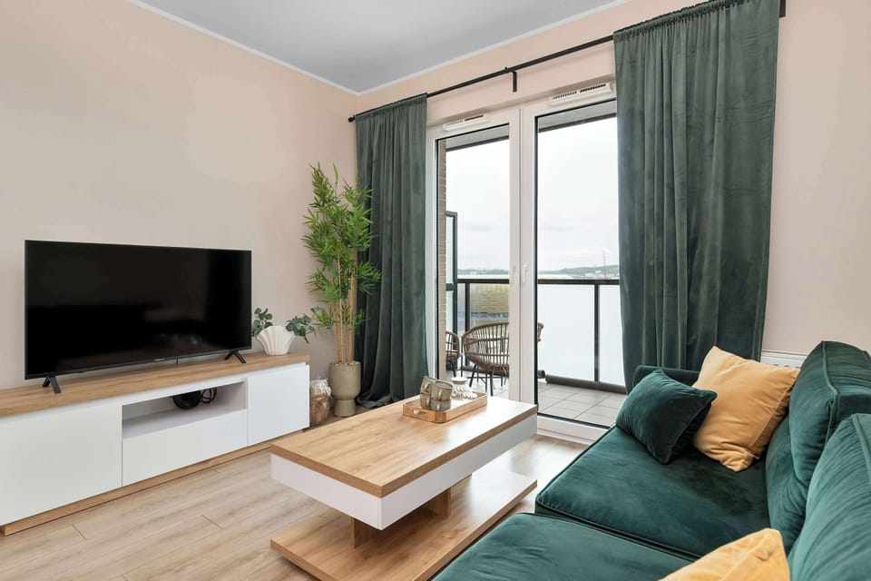 Another view of the living room showcases the television mounted on a white media console across from the plush emerald sofa. Sunlight streams in through the balcony door, illuminating the open layout.