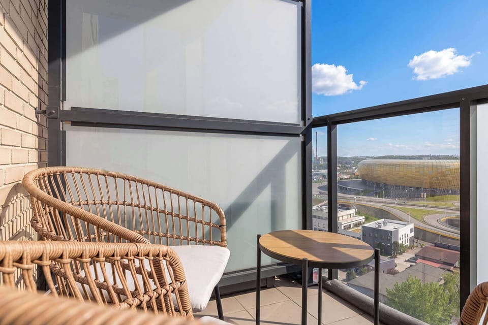 The balcony is furnished with stylish wicker chairs and a small round table, offering a private outdoor retreat. Beyond the glass railing, the surrounding neighborhood is visible under a clear blue sky.