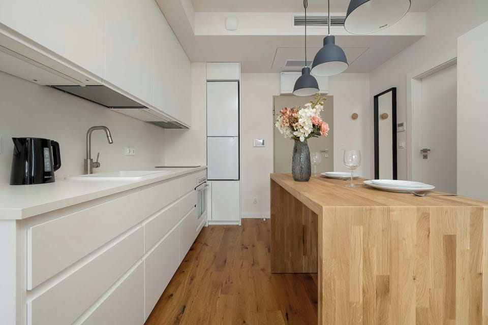 A sleek, minimalist white kitchenett corridor featuring integrated appliances and a long countertop. A wooden island stands opposite, offering a perfect spot for meal preparation or casual dining.