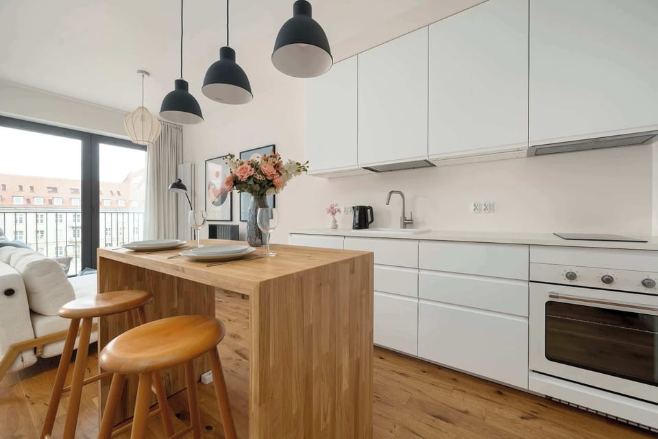A modern open-plan kitchen showcasing a beautiful wooden kitchen island with matching wooden bar stools. White minimalist cabinetry and contemporary pendant lights complete the sleek look of the dining area.