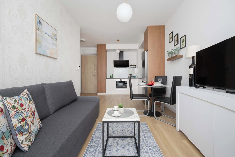 A modern living room connected to a compact white kitchen. Decorative pillows and light wooden flooring add warmth to the space.