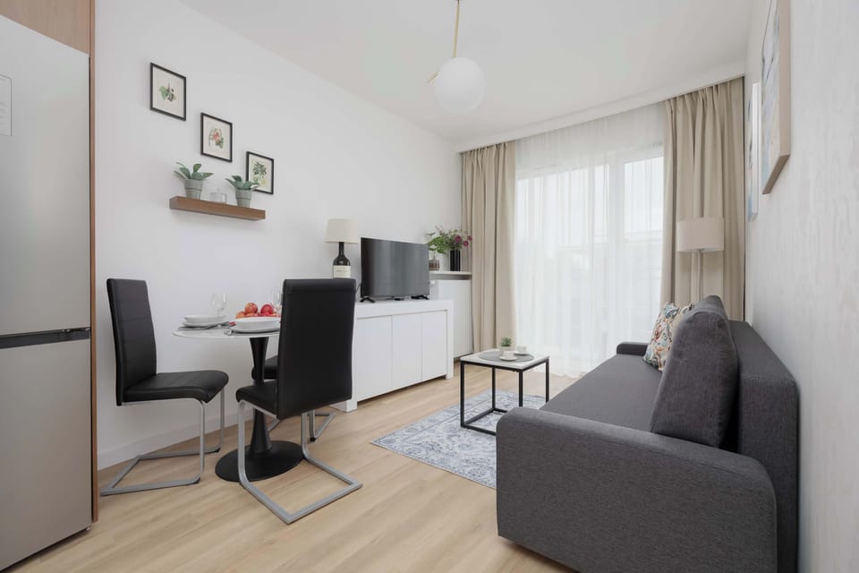 A bright living room with a gray sofa, TV, and large windows covered with light curtains. The modern interior feels cozy and spacious.