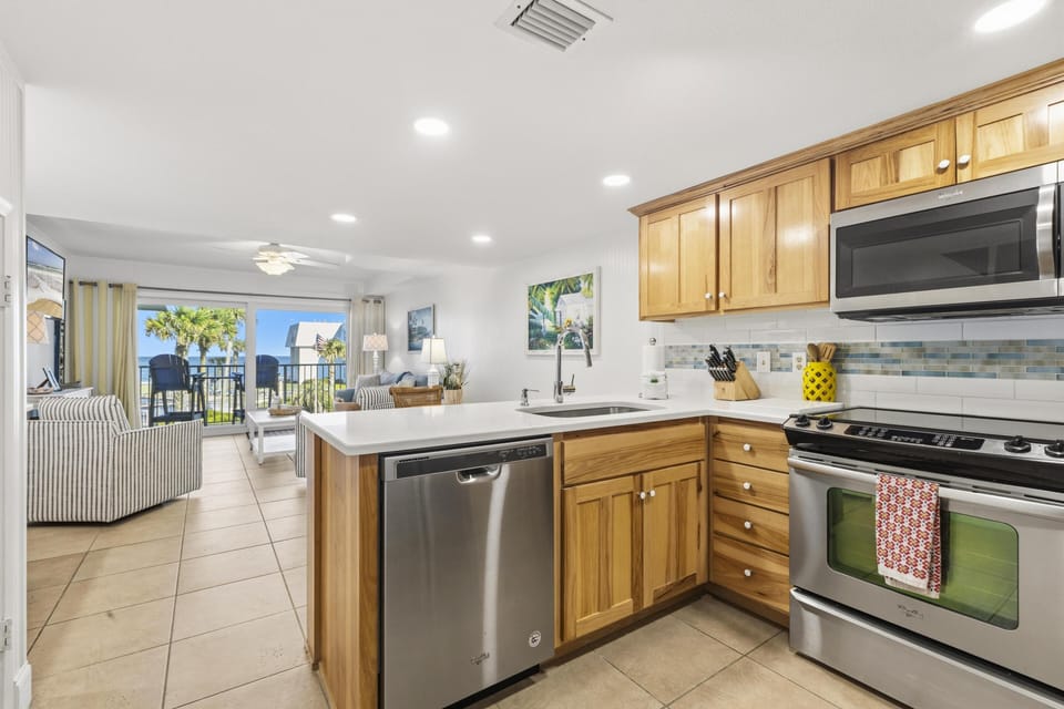 Stainless steel appliances complement honey wood cabinets and a white quartz countertop in the kitchen with tiled floor.