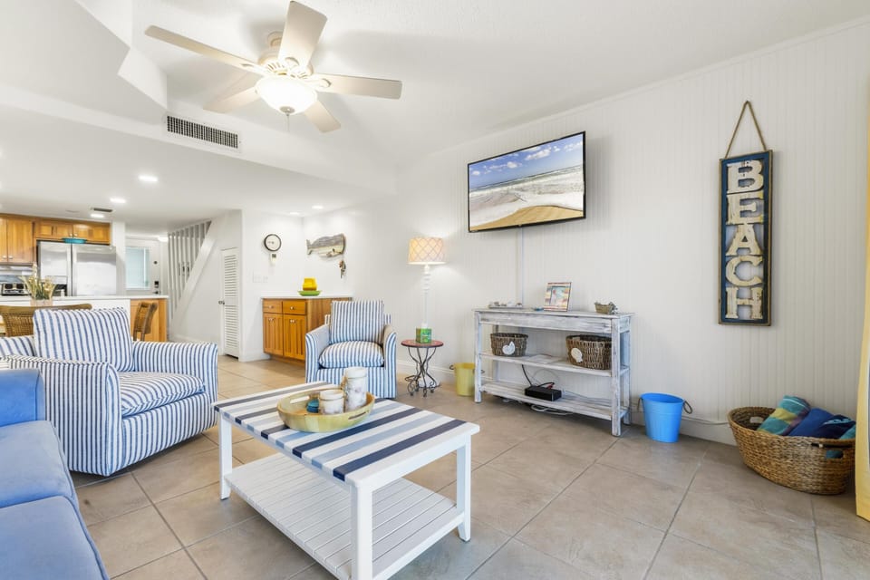 TV mounted above distressed white console with woven baskets, wicker basket of towels, and ceramic table lamp.