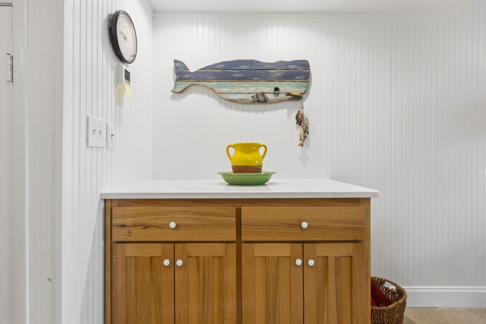Natural wood cabinet topped with white countertop, accented by a rustic blue whale wall decor and yellow vase.