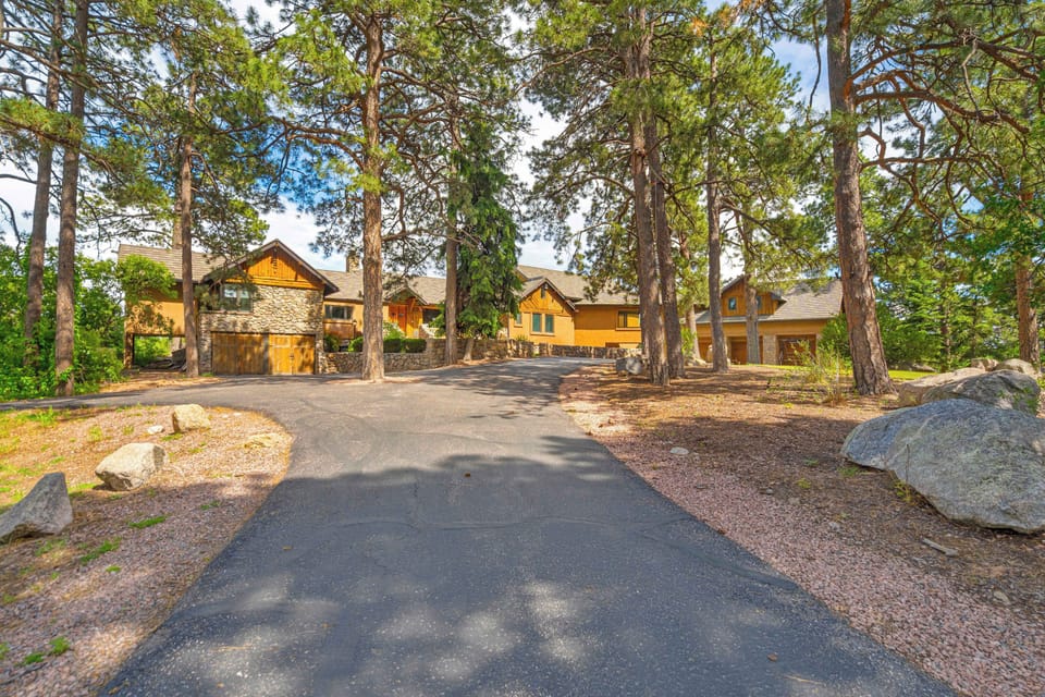 The long private driveway winds through mature pines and scrub oak toward the lodge, with the stucco and quarry stone exterior emerging through the tree canopy and a large boulder framing the entrance.