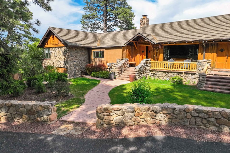 The lodge exterior showcases a warm mix of stucco, quarry stone, and natural wood trim beneath a standing seam metal roof, with manicured landscaping and a flagstone staircase leading to the main entrance.