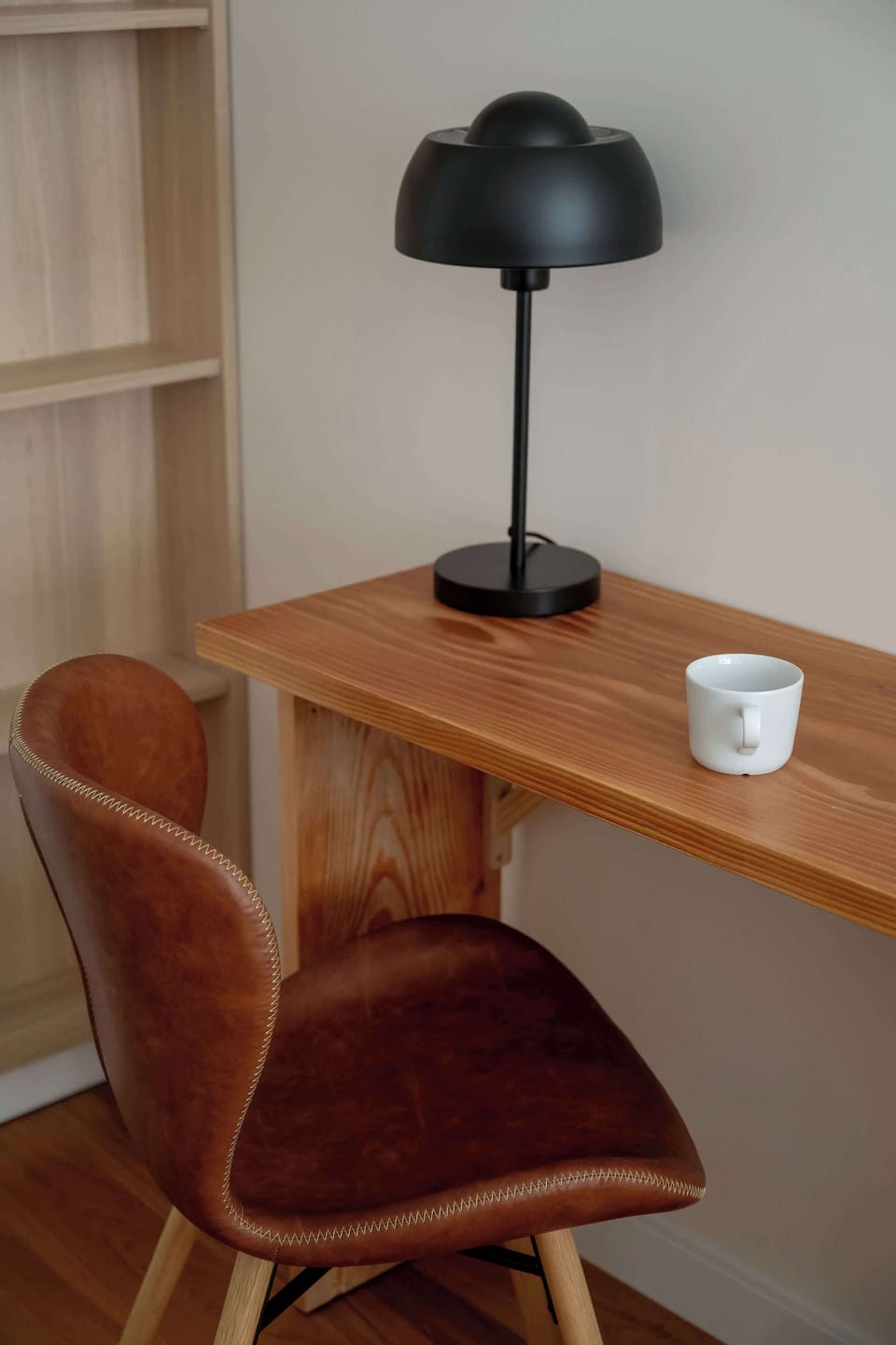 A compact workspace with a wooden desk, chair, and elegant black lamp. The open doorway leads directly toward the bedroom area.