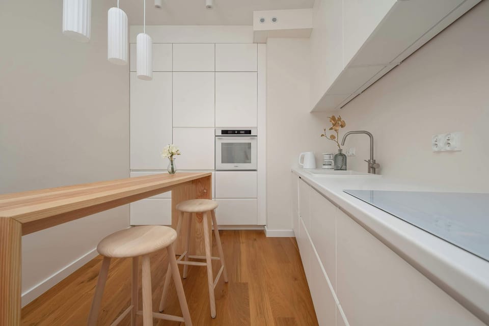 A modern kitchenette with a long breakfast bar and minimalist white cabinetry. Wooden flooring and stylish pendant lights give the interior a contemporary character.