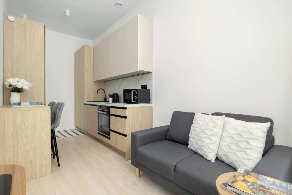 A bright studio living area with a grey sofa, a small coffee table, and large windows letting in plenty of natural light.