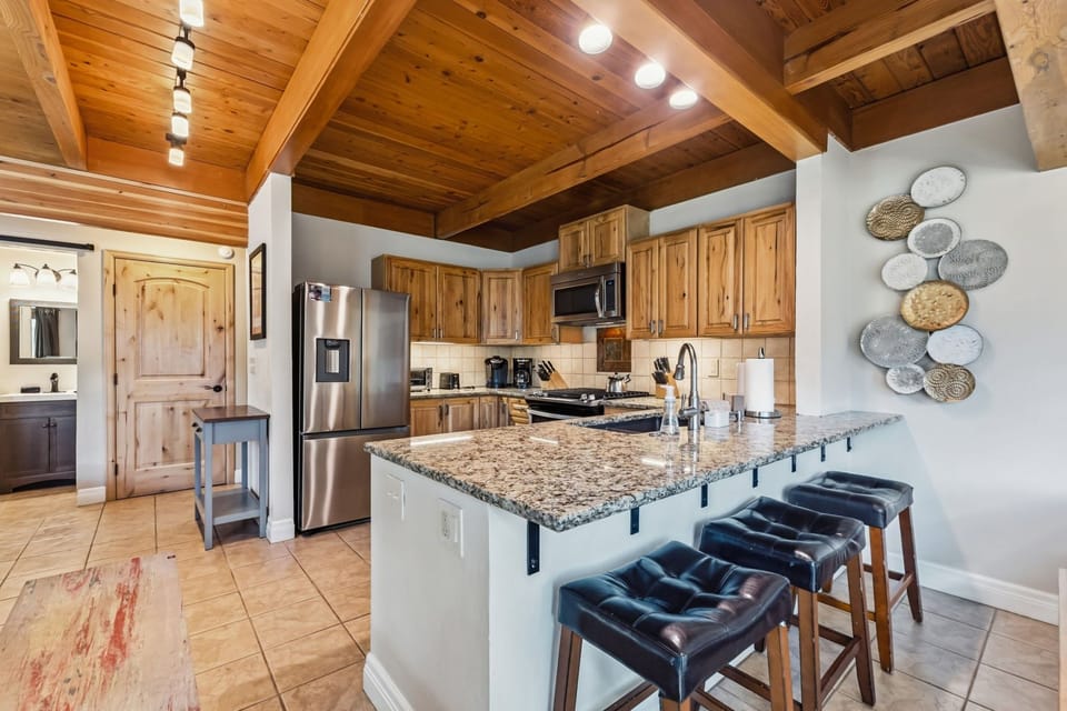 Your fully-equipped kitchen features granite counters, stainless appliances, and a cozy breakfast bar under warm wooden beams—ready for morning coffee or evening meals together.