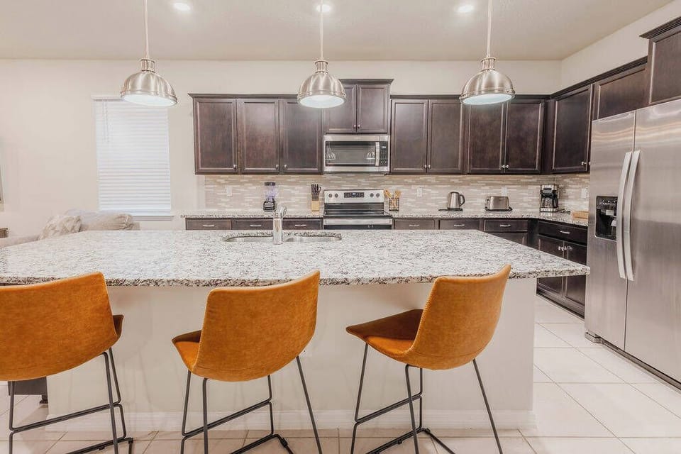 The sleek kitchen boasts rich dark wood cabinetry and stunning granite countertops for a warm, stylish vibe. Three chic rust-colored bar stools invite casual dining at the spacious island, lit by modern pendant lights.