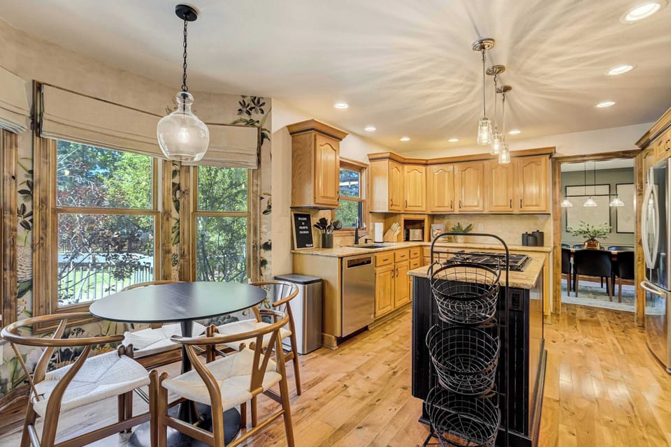 The kitchen opens into a sun-filled dining nook with a round table, woven chairs, and large windows on every side. A central island with a gas cooktop and pendant lighting keeps the workspace bright from every angle.
