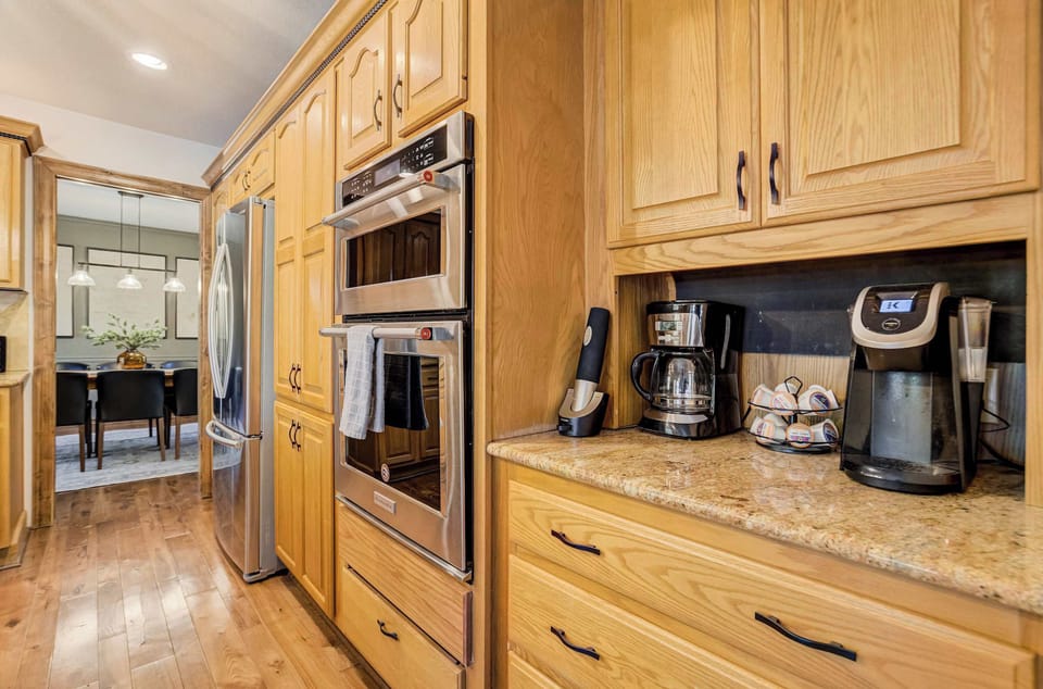 The kitchen opens toward the dining room with a round table, woven chairs, and large windows flooding the space with natural light. A central island with a gas cooktop and pendant lighting keeps the workspace bright from every angle.