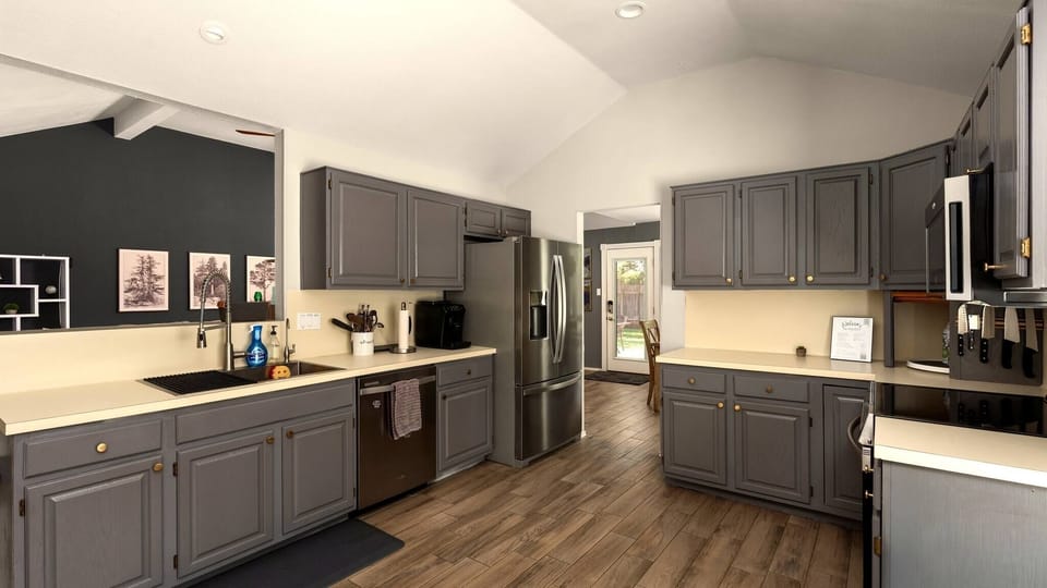 Spacious kitchen with vaulted ceilings, grey shaker-style cabinets, stainless steel French door refrigerator, dishwasher, and warm wood-look flooring — a practical, well-laid-out cooking space with room to move and everything close at hand.