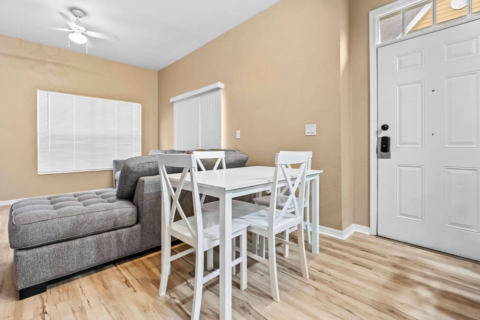 Experience a cozy atmosphere with a soft gray sectional and a bright white dining set, ideal for shared meals. The warm beige walls and natural light create a welcoming vibe, making you feel right at home.
