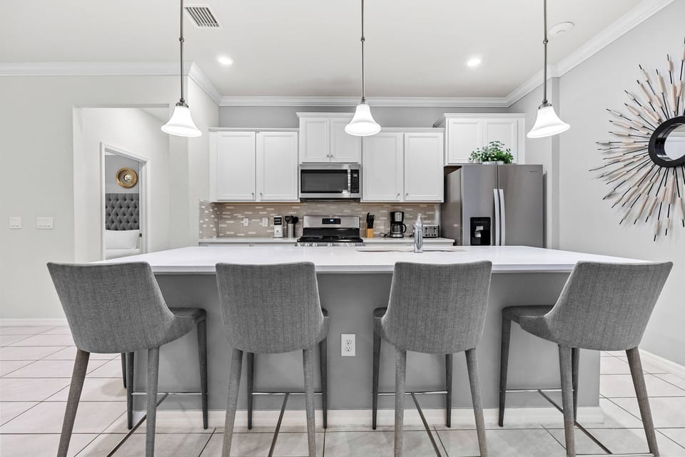 The sleek kitchen boasts a spacious island with modern gray bar stools, ideal for casual dining or morning coffee, while bright white cabinetry and elegant pendant lights create a warm and inviting atmosphere for guests to relax.