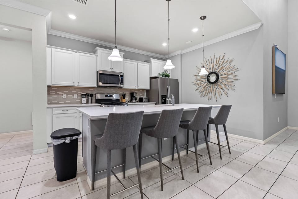 Welcome to this stylish kitchen, featuring sleek white cabinetry and a spacious island with elegant gray stone. The contemporary pendant lighting and chic circular mirror create an inviting space for gathering and dining together.
