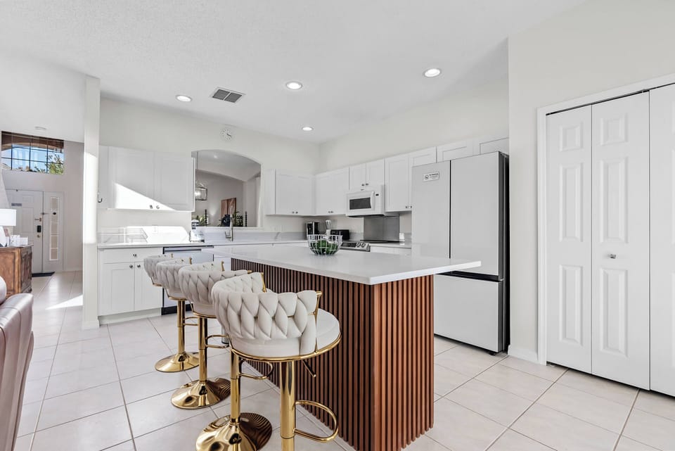 This stylish kitchen features bright white cabinetry and a large island with rich wooden accents and cozy bar stools, all bathed in natural light, making it an inviting spot for casual meals or enjoying your morning coffee.
