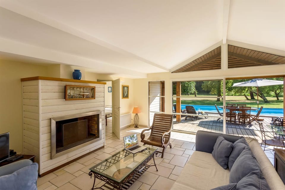 Living room with a fireplace and glass coffee table — terrace doors open wide to the pool and garden beyond.