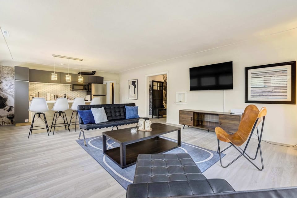 The open-concept living area features blue upholstered seating, a leather butterfly chair, dark coffee table, wall-mounted Smart TV, and a blue area rug, all flowing seamlessly into the kitchen space beyond.