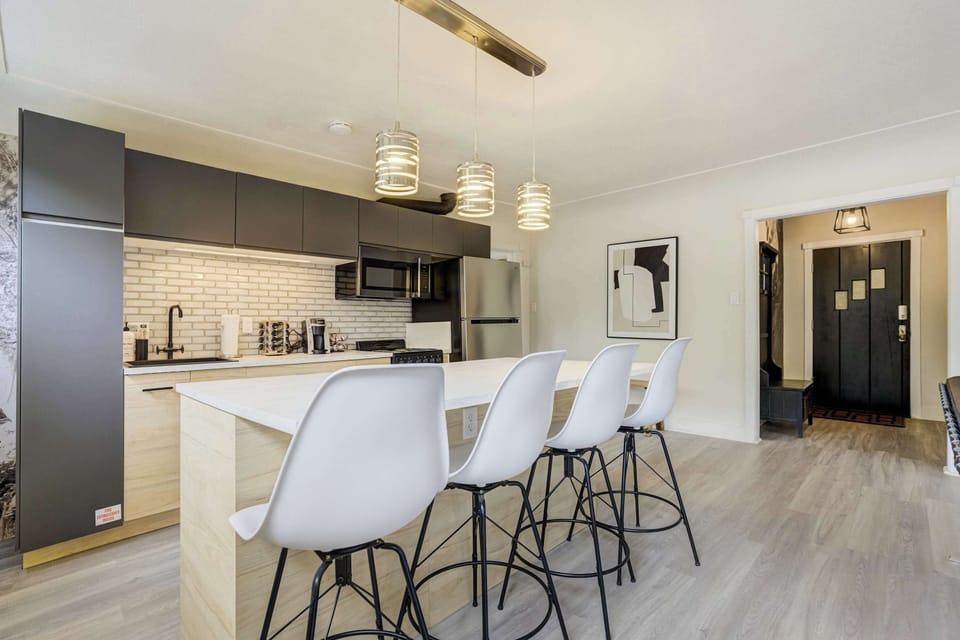 The kitchen island with white bar stools for three sits at the center of the fully stocked kitchen, perfectly positioned between the gas range, professional-grade appliances, and the open living area beyond.