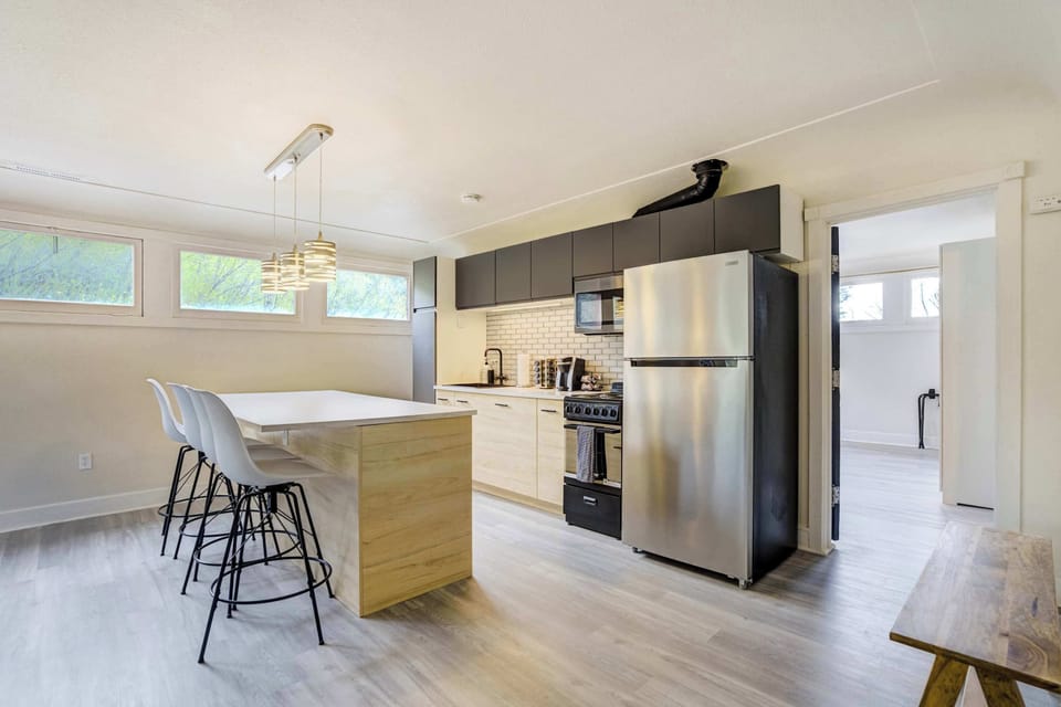 The fully stocked kitchen features a freestanding island, dark upper cabinetry, professional-grade stainless steel refrigerator, and a complimentary coffee station, with the bedroom visible through the doorway beyond.