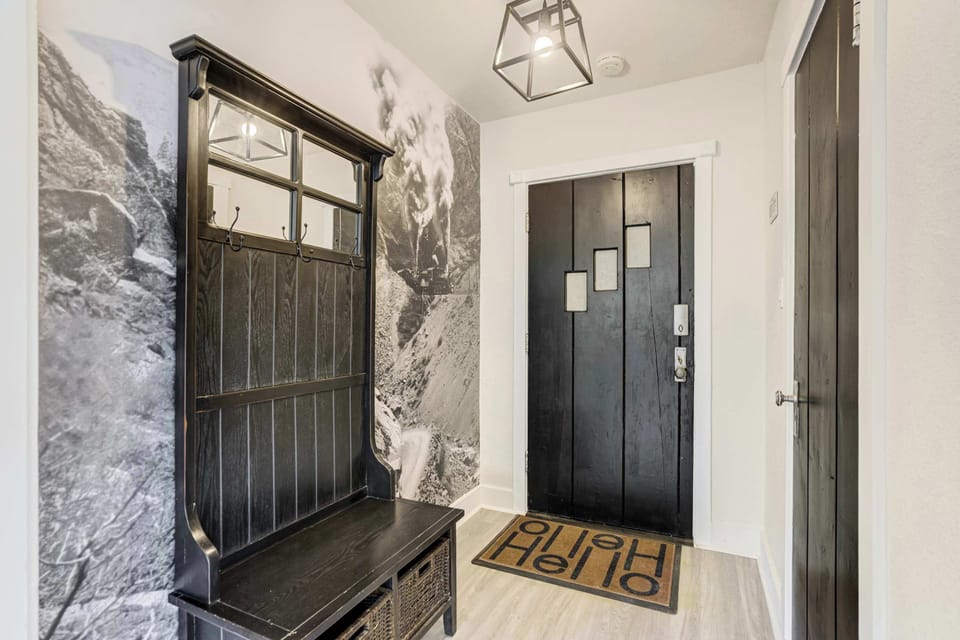 The entry foyer makes a strong first impression with a full-height black hall tree with mirror, coat hooks, and wicker storage, a cage-style pendant light, bold mountain mural wallpaper, and a black planked front door.
