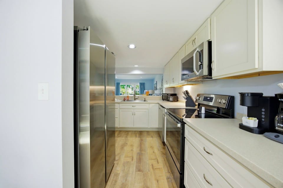 Creamy white cabinetry and brushed metal appliances create a bright, efficient space for morning coffee. Smooth stone counters meet honeyed wood floors, leading the eye toward a glimpse of garden greenery through the pass-through.