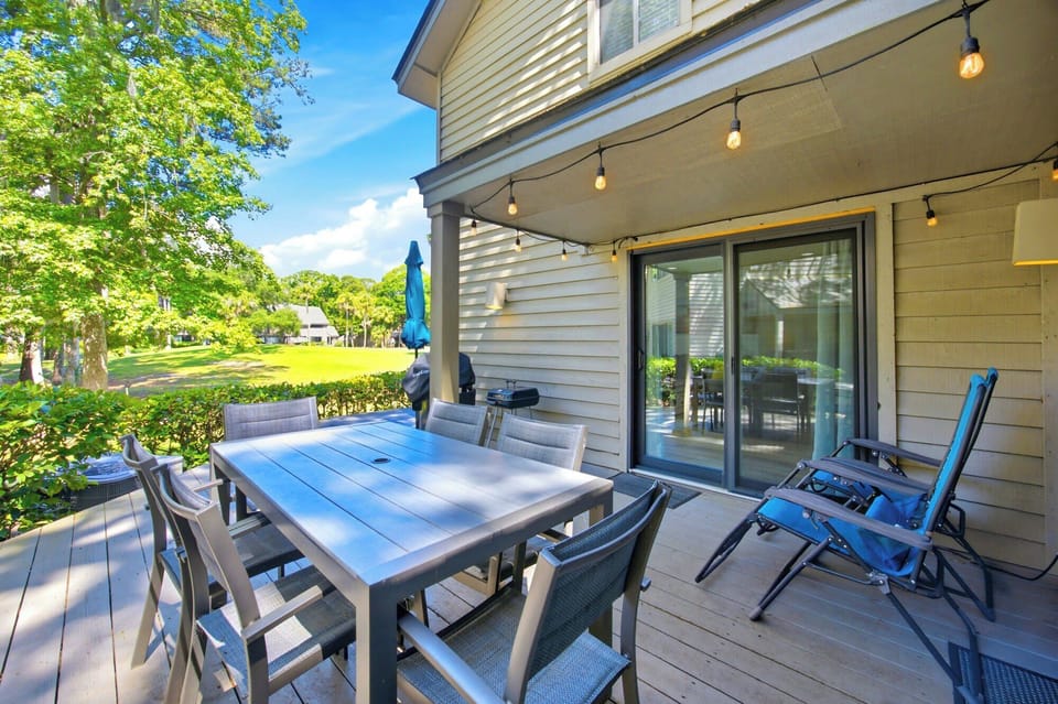 Golden afternoon rays wash over this spacious wooden deck, where string lights hang ready for a twilight dinner. Lush green lawns stretch out beyond the dining table, offering a peaceful backdrop for your morning coffee or an evening barbecue.