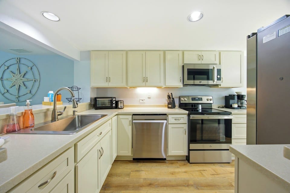 Stainless steel appliances glint under bright recessed lighting, creating a sleek contrast against creamy cabinetry. Wide plank flooring adds a grounded, organic feel to this modern culinary space, perfect for slow mornings and shared meals.