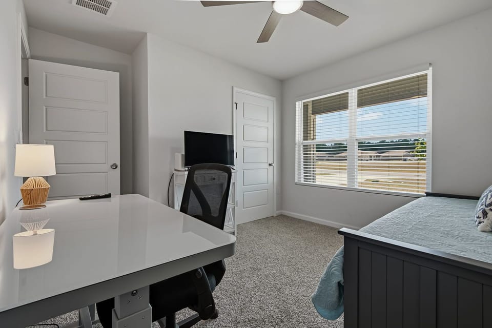 Bedroom with a twin bed and light blue blanket, white desk with a black ergonomic chair, wall-mounted TV, and a lamp
