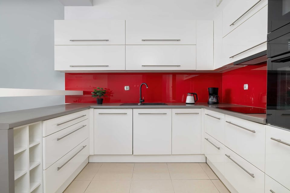 A sleek white kitchen with glossy cabinets and a vivid red backsplash adding a bold accent. Modern appliances and clean lines create a contemporary cooking space.
