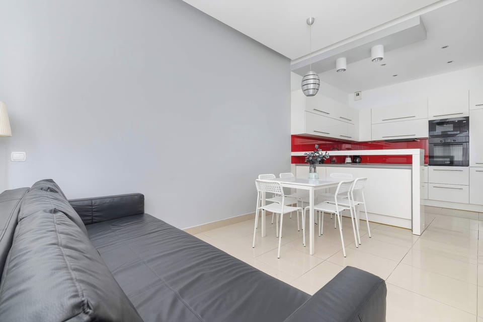 A modern studio interior with a gray sofa facing a white kitchen and dining corner. Minimalist furniture and soft lighting enhance the clean design.