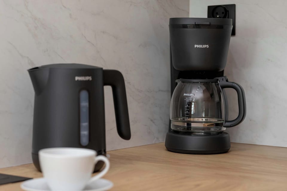A close-up of kitchen amenities including a matte black electric kettle and a matching drip coffee maker sitting on a wooden surface. A white ceramic coffee cup and saucer rest neatly in the foreground, ready for use.