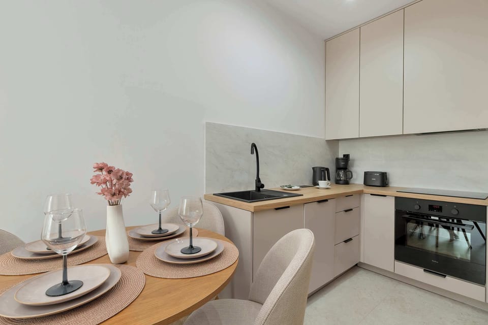 A compact, modern kitchenette corner featuring light beige cabinetry, a wooden countertop, and an integrated oven. A round dining table with plates, glasses, and a small vase with flowers is set for a meal.