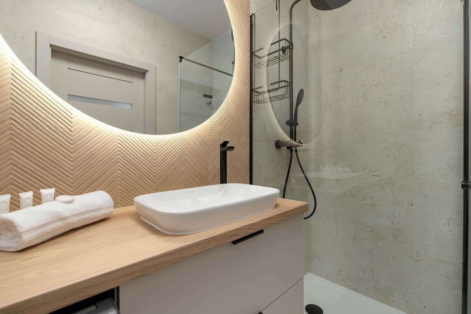A close-up view of a bathroom vanity featuring a white vessel sink sitting on a light wood countertop. A large round mirror with integrated LED backlighting hangs on a textured, wood-paneled wall next to a glass shower screen.