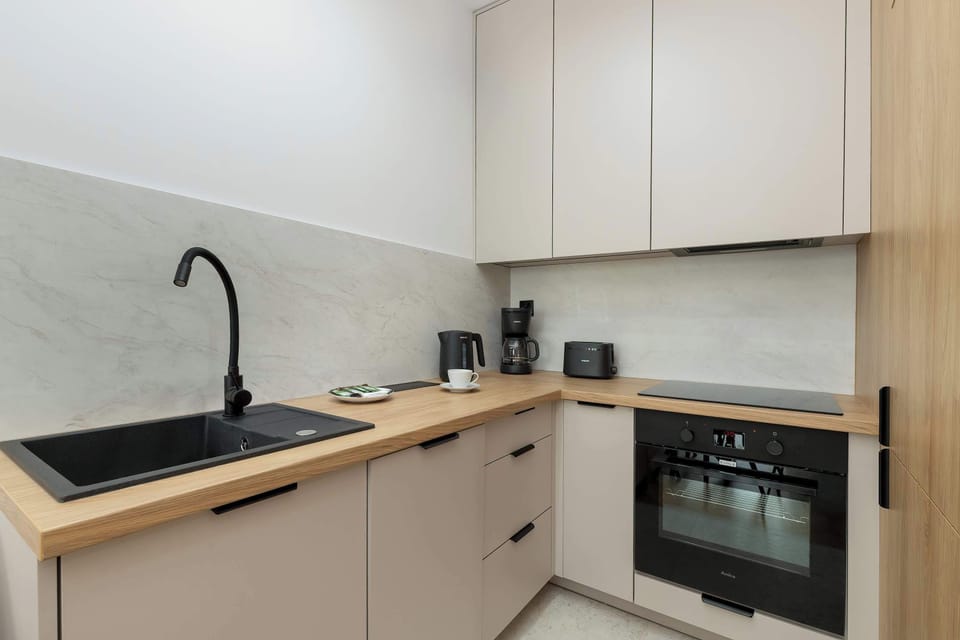 A functional L-shaped kitchenette with light-colored cabinets, a black sink with a matching faucet, and an induction cooktop. The marble-patterned backsplash adds an elegant touch to the clean and minimalist cooking space.