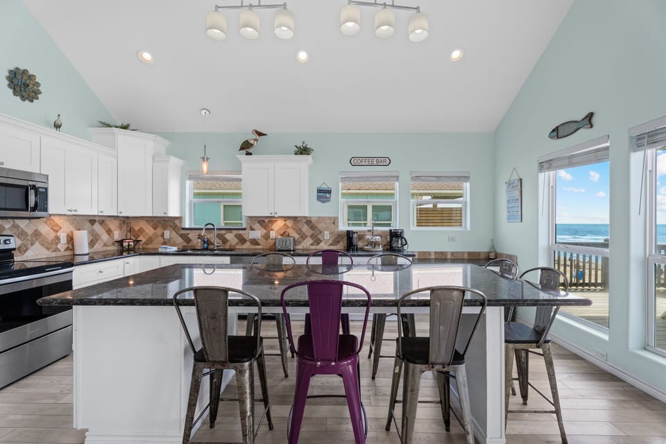 Open dining and kitchen area with large island seating, stainless steel appliances, and bright coastal décor perfect for group vacations.