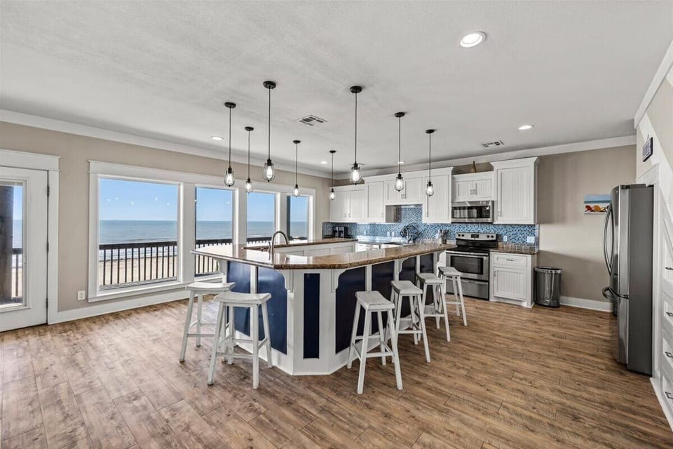 Bright beachfront kitchen with bar seating, wood floors, modern lighting and open views to the outdoor balcony and Gulf Coast scenery.